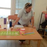 Zdravstvena delavka stoji za mizo z zobozdravstvenimi pripomočki in se pogovarja s starejšo gospo. Na mizi so modeli zob, zobne ščetke in informativne zloženke.