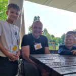 Nasmejani deček stoji ob odraslem moškem in drugem otroku, vsi nosijo obeske s slikami sadja.