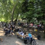Večja skupina ljudi, med njimi številni v vozičkih, sedi in stoji pod drevesi v parku, ob mizah z osvežilno postrežbo.