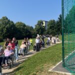 Dolga kolona stanovalcev v invalidskih vozičkih in spremljevalcev hodi po poti mimo športnega igrišča na sončen dan.