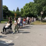 Skupina starejših oseb v vozičkih in njihovih spremljevalcev prečka cesto na prehodu za pešce, obdani z zelenjem.