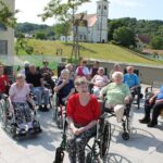 Večja skupina starejših oseb sedi zunaj na terasi doma starejših občanov. V ozadju je cerkev na hribčku in okoliške hiše. Osebe spremljajo dogajanje pred seboj.