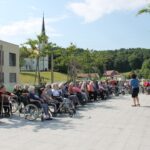 Pogled iz daljave na dogodek na terasi doma za starejše. Ženska z mikrofonom stoji pred dolgo vrsto obiskovalcev, ki sedijo in poslušajo. V ozadju je cerkev in zeleno podeželje.