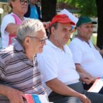 Trije moški srednjih let in starejši sedijo na klopi zunaj, držijo slovenske zastavice, eden nosi rdečo kapo z napisom »Ptujčani«, v ozadju spremljevalno osebje.
