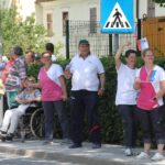Skupina ljudi, med njimi stanovalci doma in osebje, stoji ob cesti z majhnimi slovenskimi zastavicami, nekateri mahajo, eden izmed stanovalcev sedi v invalidskem vozičku.