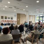 Otroci v oranžnih kapah nastopajo pred starejšimi osebami, ki jih gledajo iz občinstva. Dvorana je svetla in prostorna z velikimi okni in slikami na stenah.