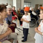 Dva otroka v oranžnih kapah se pogovarjata z dvema starejšima ženskama, ki sedita. V ozadju so drugi stanovalci, nekateri v invalidskih vozičkih.
