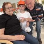 Deček v oranžni kapi stoji med nasmejano starejšo gospo in gospodom. Gospa sedi in drži telefon, gospod pa otroka drži za rame.