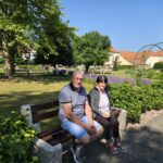 Starejši moški in ženska sedita na klopi v urejenem vrtu z nasadom sivke in pogledom na stavbo doma.
