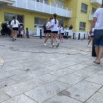 Skupina deklet v belih majicah in črnih kratkih hlačah izvaja plesno točko pred občinstvom na dvorišču doma za starejše.