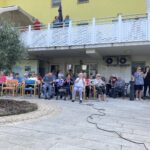 Gledalci, večinoma starejši, sedijo na dvorišču doma za starejše in spremljajo nastop, nekateri sedijo v invalidskih vozičkih.