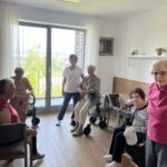 Šest starejših žensk in ena zaposlena stojijo ali sedijo v svetli sobi doma za starejše. V ozadju je balkon s pogledom na zunanji svet.
