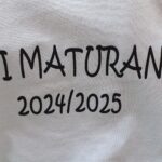 Povečan izrez bele majice z napisom “Mini maturanti 2024/2025” v črni pisavi, kot del otroškega nastopa ali praznovanja zaključka vrtca.