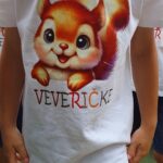 Majica z velikim tiskanim motivom nasmejane risane veverice in spodnjim napisom “Veveričke” v barviti pisavi, ki označuje skupino otrok.