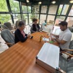 Strokovnjak v beli majici sedi za veliko leseno mizo v svetli sobi z velikimi steklenimi stenami, ob mizi orodje in kovček, nasproti njemu sedita dve starejši ženski.