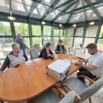 Perspektiva od zadaj: strokovnjak sedi za mizo z orodjem in kovčkom, pred njim sta dve starejši ženski v invalidskih vozičkih, ambient svetel s steklenim stropom.