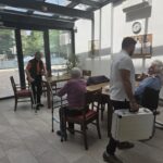 Strokovnjak z belim kovčkom v roki stoji ob mizi, pred njim ženska v vozičku drži žlico, v ozadju dve ženski z berglami in pohodno hoduljo.