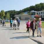 Skupina starejših oseb hodi po cesti pred novo stavbo, nekatere uporabljajo bergle in hodulje, v ozadju se vidi znak avtobusne postaje.