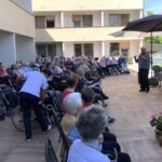 Moški v sivem puloverju govori na prireditvi doma starejših, stanovalci in osebje pa ga poslušajo s terase.