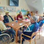 Skupina starejših moških sedi okoli lesene mize v notranjem prostoru, pogovarjajo se in pijejo kavo. Eden izmed njih je v invalidskem vozičku.