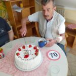 Starejši moški v beli majici in brezrokavniku sedi za mizo ter reže torto, okrašeno z jagodami in številko 70. Ob njem so skodelica, kozarec in znak s številko 70.