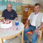 Dva moška sedita za okroglo mizo, na kateri je rojstnodnevna torta z jagodami in napisom 70. Eden izmed njiju nosi majico s potiskom, drugi telovnik in kavbojke.