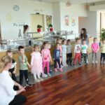 Skupina otrok iz vrtca stoji v vrsti pred jedilnico doma za starejše in nastopa za stanovalce. Ob strani jih spremljata dve vzgojiteljici.