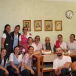 Skupina zaposlenih v domu za starejše skupaj s stanovalcem praznuje rojstni dan. V ospredju na mizi je torta, vsi se smejijo v kamero.