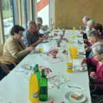 Skupina stanovalcev doma za starejše sedi okoli dolge mize, okrašene z rožami in prtički, ter uživa ob pijači in kosih torte.