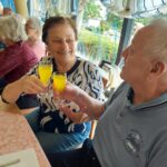 Starejši moški in ženska si z nasmehom nazdravljata s kozarcema soka ob pogrnjeni mizi.