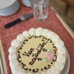 Praznična torta z napisom »Vse najboljše!«, obdana s smetano in okrašena s cvetovi iz kreme, pripravljena za razrez na vozičku poleg krožnikov in noža.