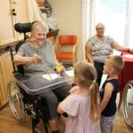 Deklica iz vrtca podarja rožico starejši gospe v invalidskem vozičku, medtem ko jo spremljata še dva otroka. V ozadju sedi še ena starejša gospa.