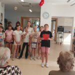 Skupina deklic in dečkov stoji v vrsti in nastopa pred stanovalci doma, ena deklica bere z rdečega listka.