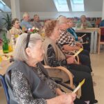 Stanovalci doma sede ob praznično okrašeni mizi in spremljajo nastop otrok, v ozadju knjižne police.