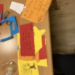 Na mizi ležijo barvni izrezani listi z ročno napisanimi sporočili in risbami. Vidna je tudi škarjica in prazna steklena posoda.