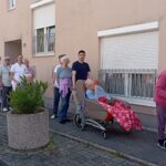 Sprehod stanovalcev doma za starejše ob stavbi doma; nekateri uporabljajo invalidske vozičke ali hodulje, osebje jih spremlja.