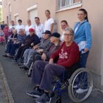 Skupina stanovalcev doma za starejše in zaposleni med postankom za fotografijo ob stavbi; več stanovalcev sedi v vozičkih, osebje stoji ob njih.