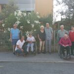 Skupinska fotografija starejših oseb ob cvetočem grmovju, z dvema osebama v invalidskih vozičkih. Ena oseba čepi ob vozičku in se nasmiha.
