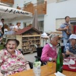 Stanovalci doma sedijo ob mizi na dvorišču, v ozadju igra harmonikar.