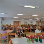 Odrasli in starejši udeleženci v domu za starejše osebe sodelujejo na pogovoru z gostujočo farmacevtko, ki sedi nasproti njih.