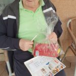 Starejši moški sedi z darilno vrečko v naročju in izpolnjenim tombola kartončkom v roki, z nasmeškom gleda v kamero.