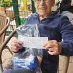 Starejša ženska sedi na stolu in drži tombola kartonček ter darilno vrečko z živili, veselo pozira za fotografijo.