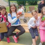 Štiri deklice sedijo na klopi na prostem in ponosno držijo darilne vrečke s sladkarijami in sokovi, v ozadju starejši moški v invalidskem vozičku.