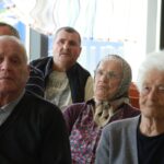Portretni pogled na več stanovalcev, ki s pozornostjo poslušajo dogajanje pred njimi. V ospredju so trije starejši posamezniki, dva moška in ena ženska z ruto.