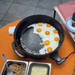 Ponev z več jajci na oko, ki se pečejo; poleg so kovinske posode s prilogami in krožniki.
