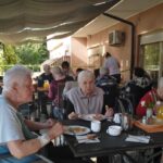 Skupina starejših oseb med zajtrkom na terasi doma starejših občanov, ob mizah s krožniki hrane, skodelicami in sokom.