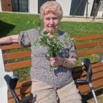Starejša ženska z berglami sedi na klopi na soncu in v rokah drži šopek svežih zelišč.