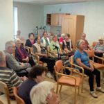 Skupina starejših oseb sedi v krogu na dogodku v skupnem prostoru doma za starejše, pozorno spremljajo program ali nastop.