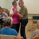 Zaposlena v domu za starejše in stanovalec skupaj plešeta, medtem ko drugi stanovalci sedijo in spremljajo dogodek.