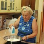 Starejša ženska v modri majici z električnim mešalnikom stepá smetano v veliki kovinski posodi na kuhinjskem pultu.